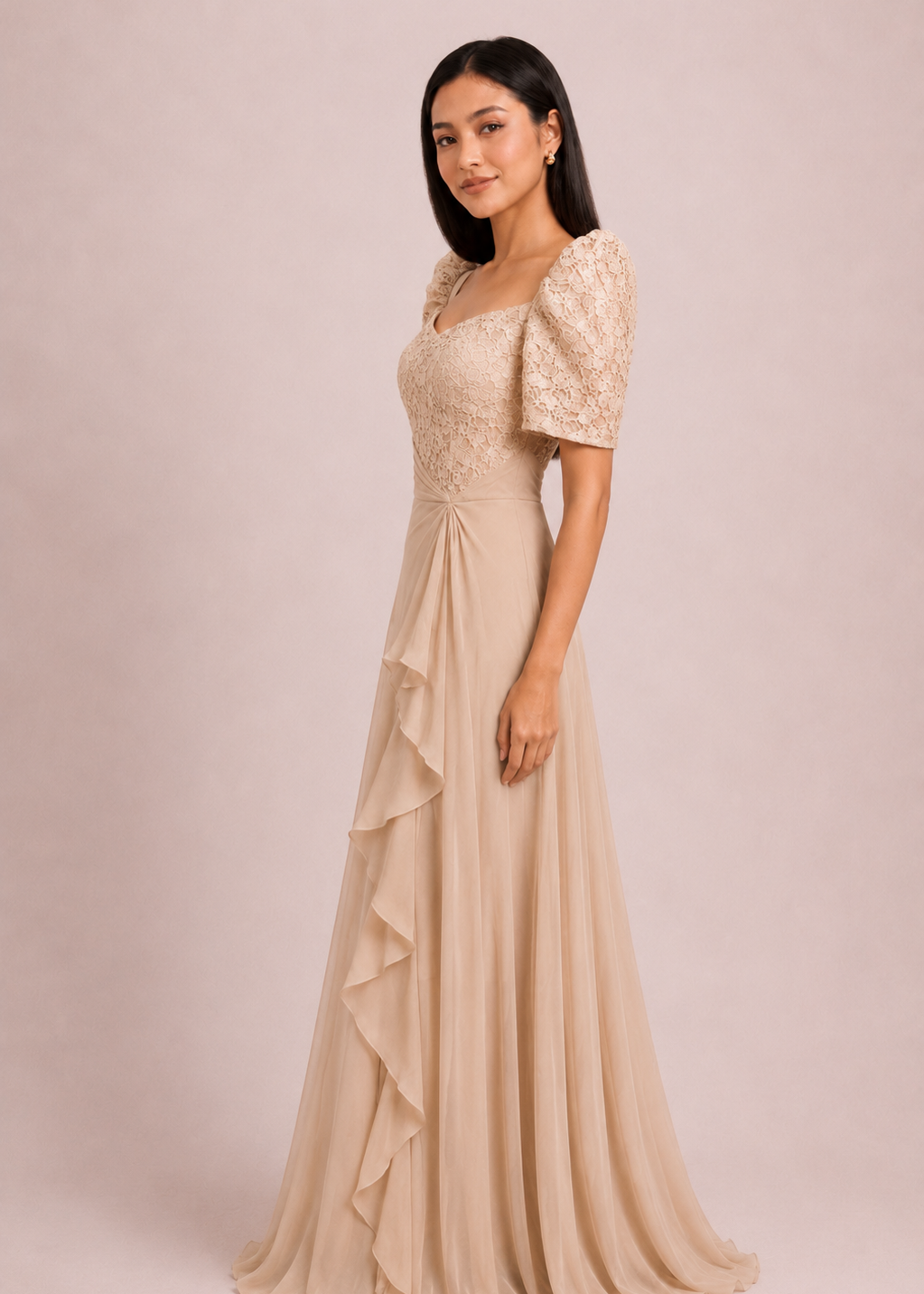 
Corazon filipiniana dress in cream pleated chiffon - A woman modeling a floor-length champagne beige evening gown with short puff sleeves, a lace bodice, and a flowing ruffled skirt, set against a solid pale pink studio background.